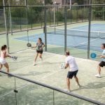Quelles sont les différences entre le padel et le tennis ? Vue en légère plongée d’un court de padel vitré et d’un court de tennis adjacent, avec quatre joueurs débutants en tenue de sport sous une lumière naturelle réaliste.