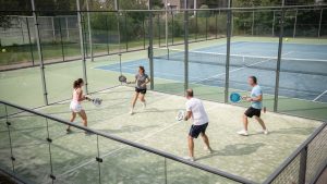 Vue en légère plongée d’un court de padel vitré et d’un court de tennis adjacent, avec quatre joueurs débutants en tenue de sport sous une lumière naturelle réaliste.