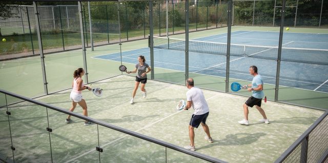 Vue en légère plongée d’un court de padel vitré et d’un court de tennis adjacent, avec quatre joueurs débutants en tenue de sport sous une lumière naturelle réaliste.