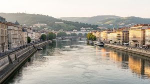 Vue urbaine réaliste de Lyon depuis les quais de la Saône, avec les immeubles anciens, la Presqu’île et les collines baignés par une douce lumière de fin de journée.